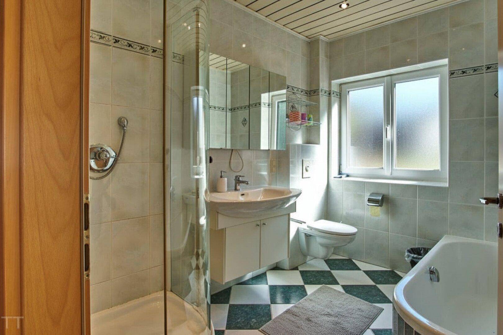 Helles Badezimmer mit Dusche, Badewanne, Fenster und kariertem Fliesenboden.