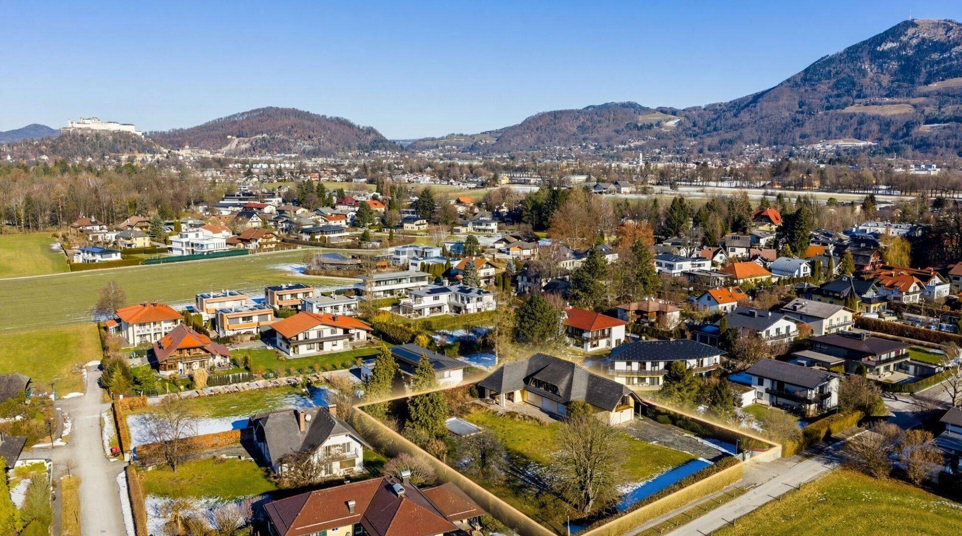 Luftaufnahme eines Wohngebiets mit vielen Häusern, grünen Flächen und Hügeln im Hintergrund.