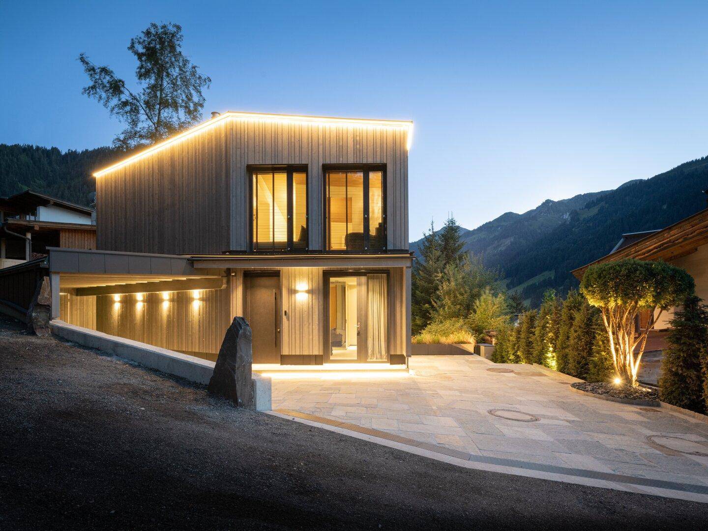 Modernes Haus bei Dämmerung mit beleuchteter Fassade und Bergblick.