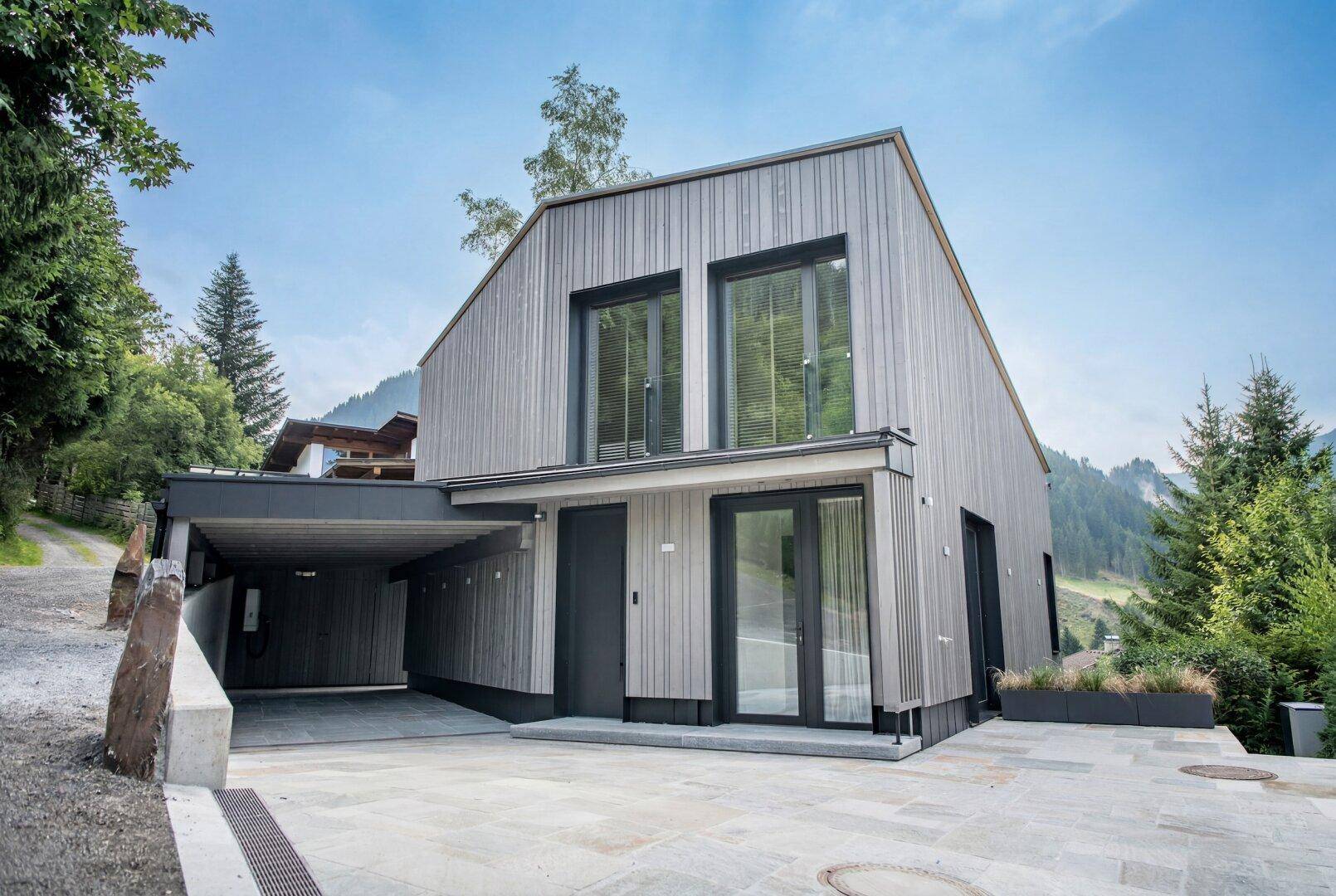 Modernes Holzhaus mit Carport und großem Vorplatz in alpiner Umgebung.
