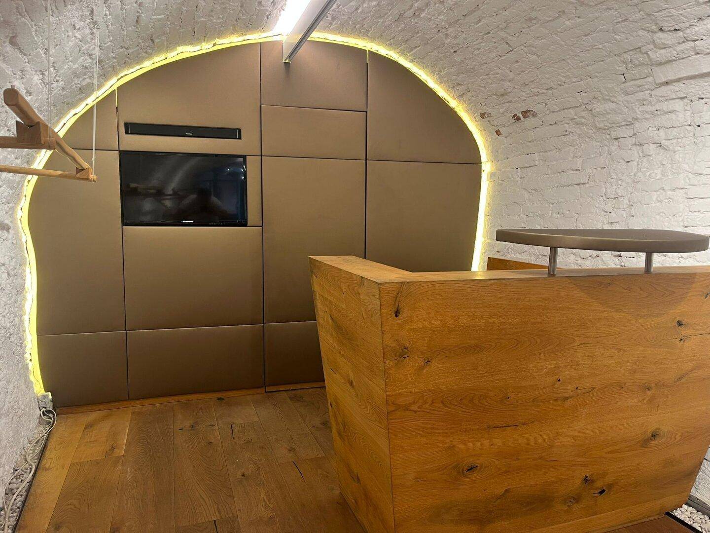Moderner Empfangsbereich mit Holztresen, TV-Bildschirm und indirekter Beleuchtung in einem Gewölberaum.