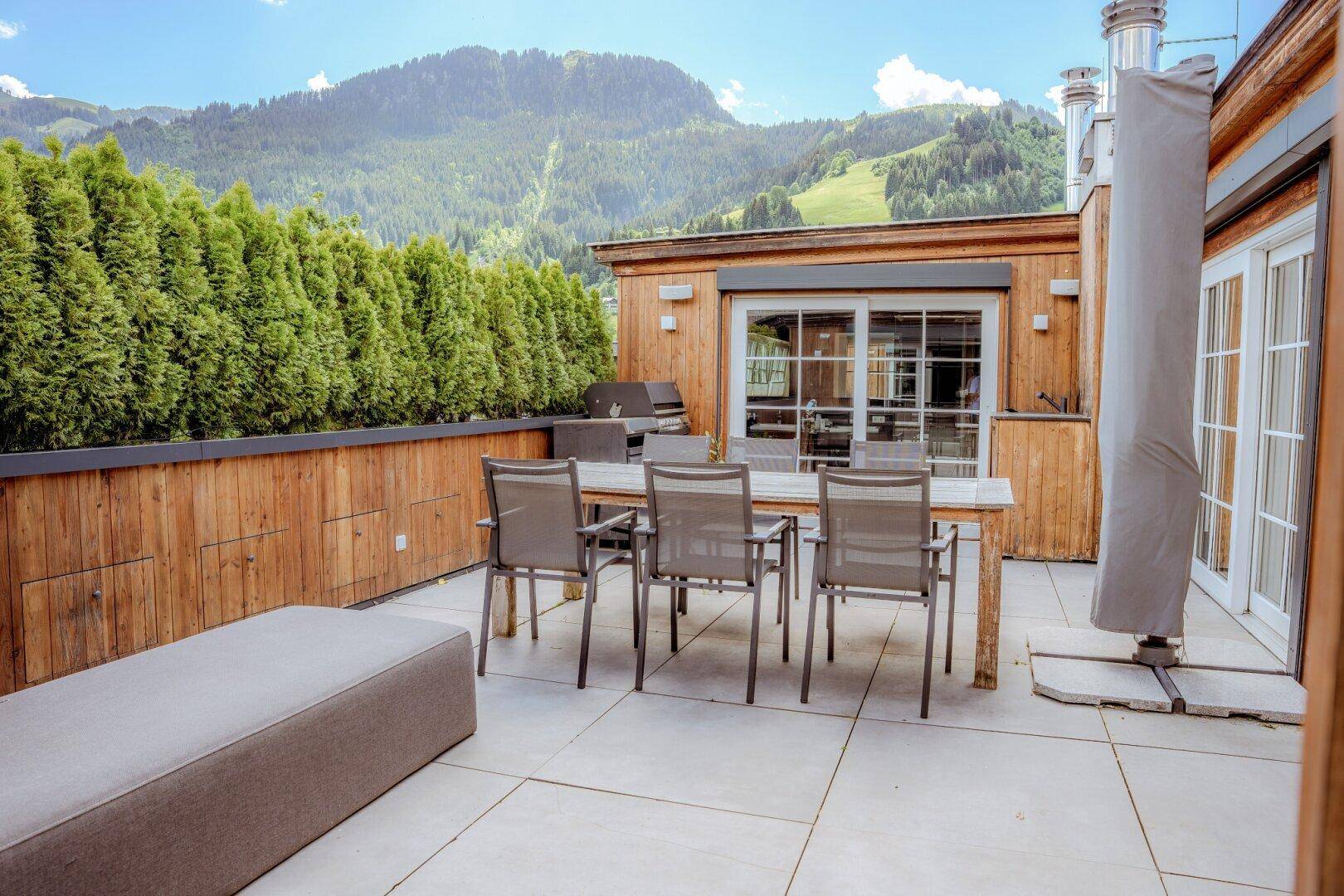 Große Dachterrasse mit Essbereich, Grill und Hecke, umgeben von einer beeindruckenden Bergkulisse.