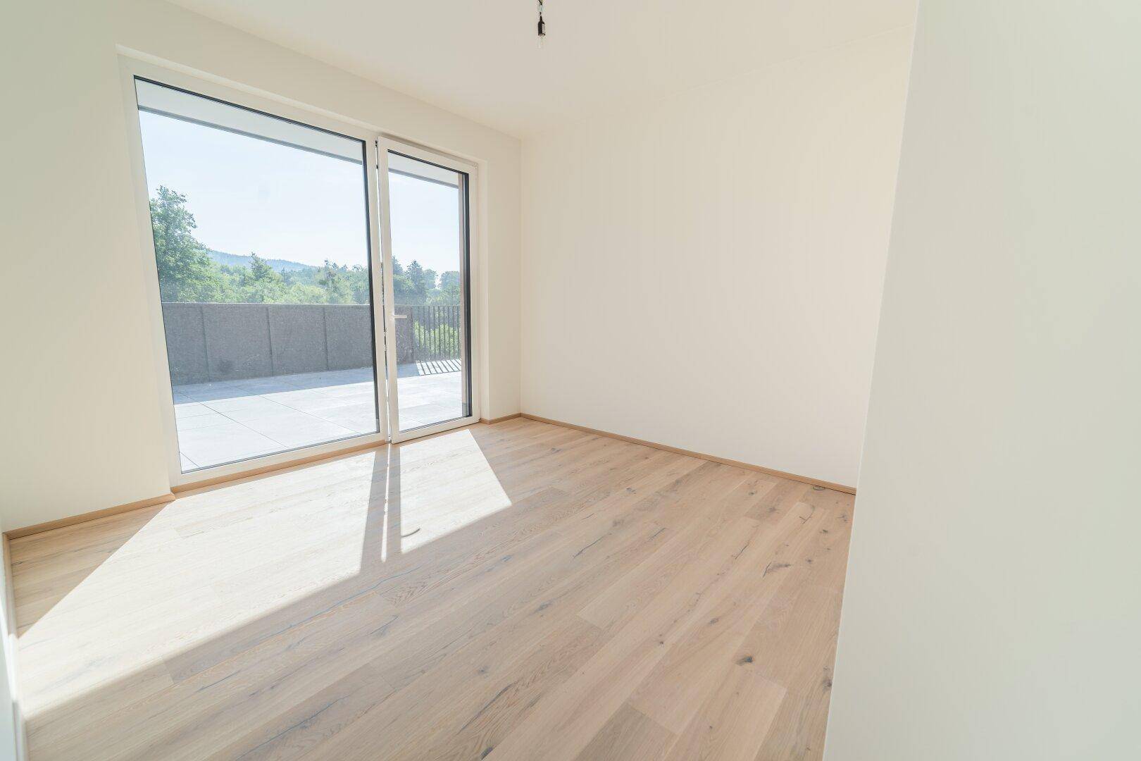 Sonniger Raum mit Holzboden und großen Fenstern, die auf eine private Terrasse führen.