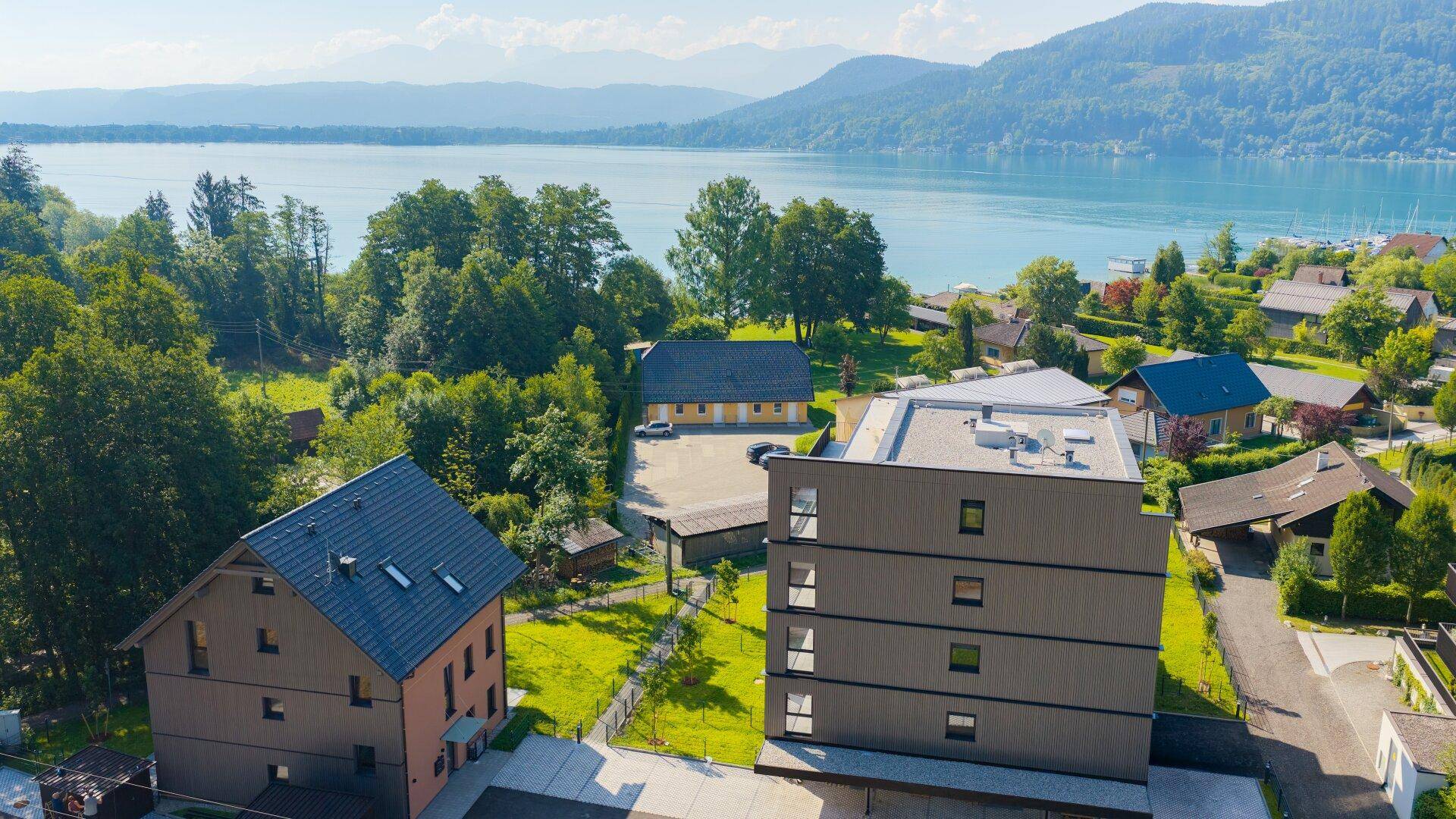Luftaufnahme von Wohngebäuden und dem Wörthersee, umgeben von bewaldeten Hügeln.