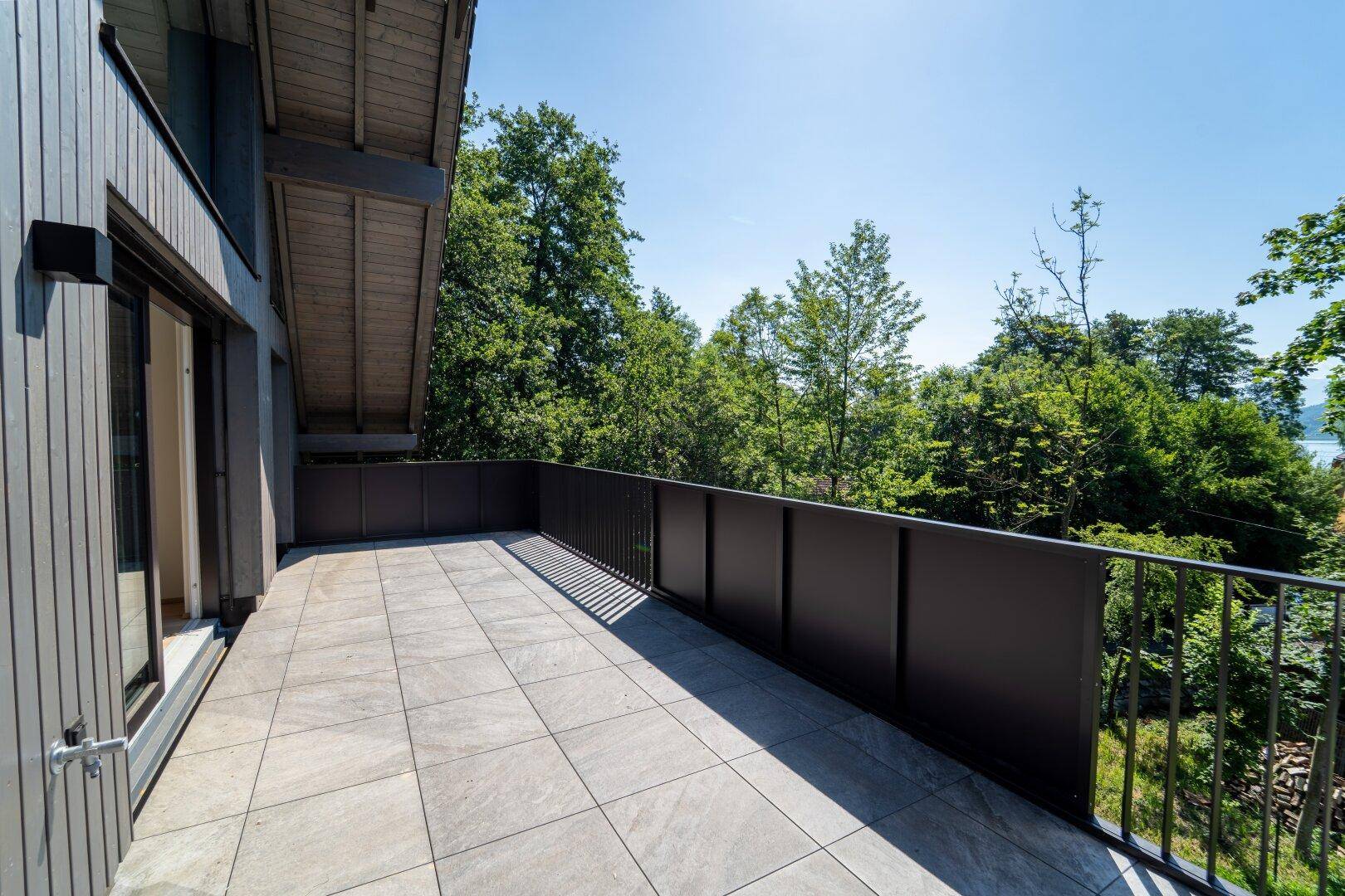 Geräumiger Balkon mit Fliesenboden und schwarzem Geländer, umgeben von Bäumen mit Seeblick.
