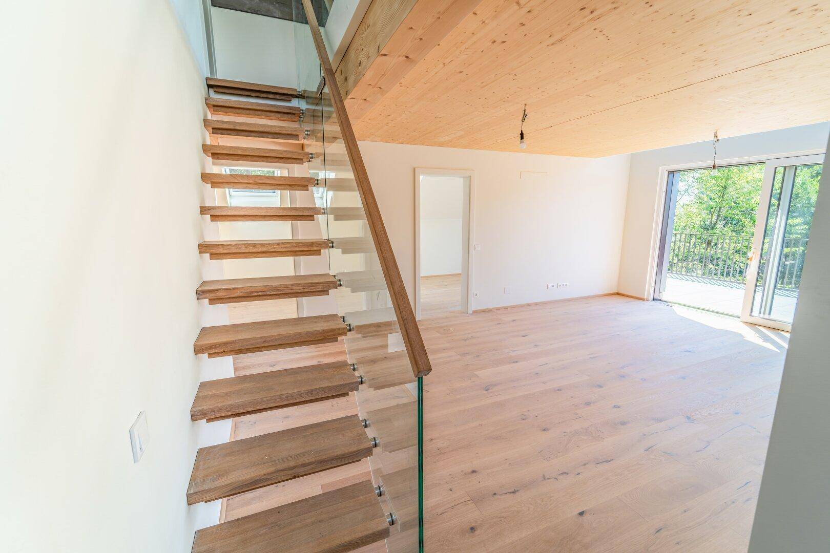 Moderne Holztreppe mit Glasgeländer in einem hellen Raum mit Holzboden und Holzdecke.