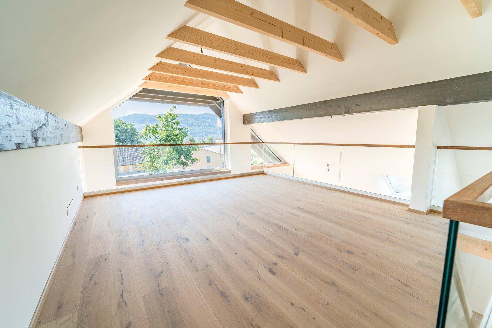 Lichtdurchflutete Galerie mit Holzboden, schrägen Decken und Panoramablick auf die umliegende Natur.