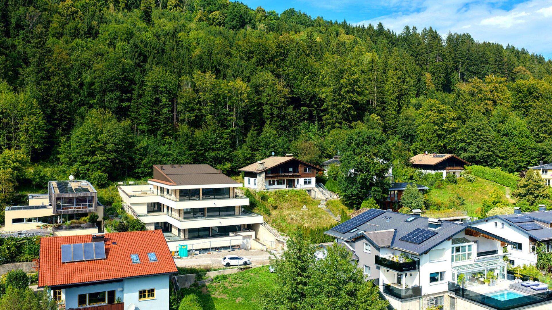 Luftaufnahme moderner Häuser am Hang mit Solaranlagen und einem Pool, eingebettet in grüne Landschaft.