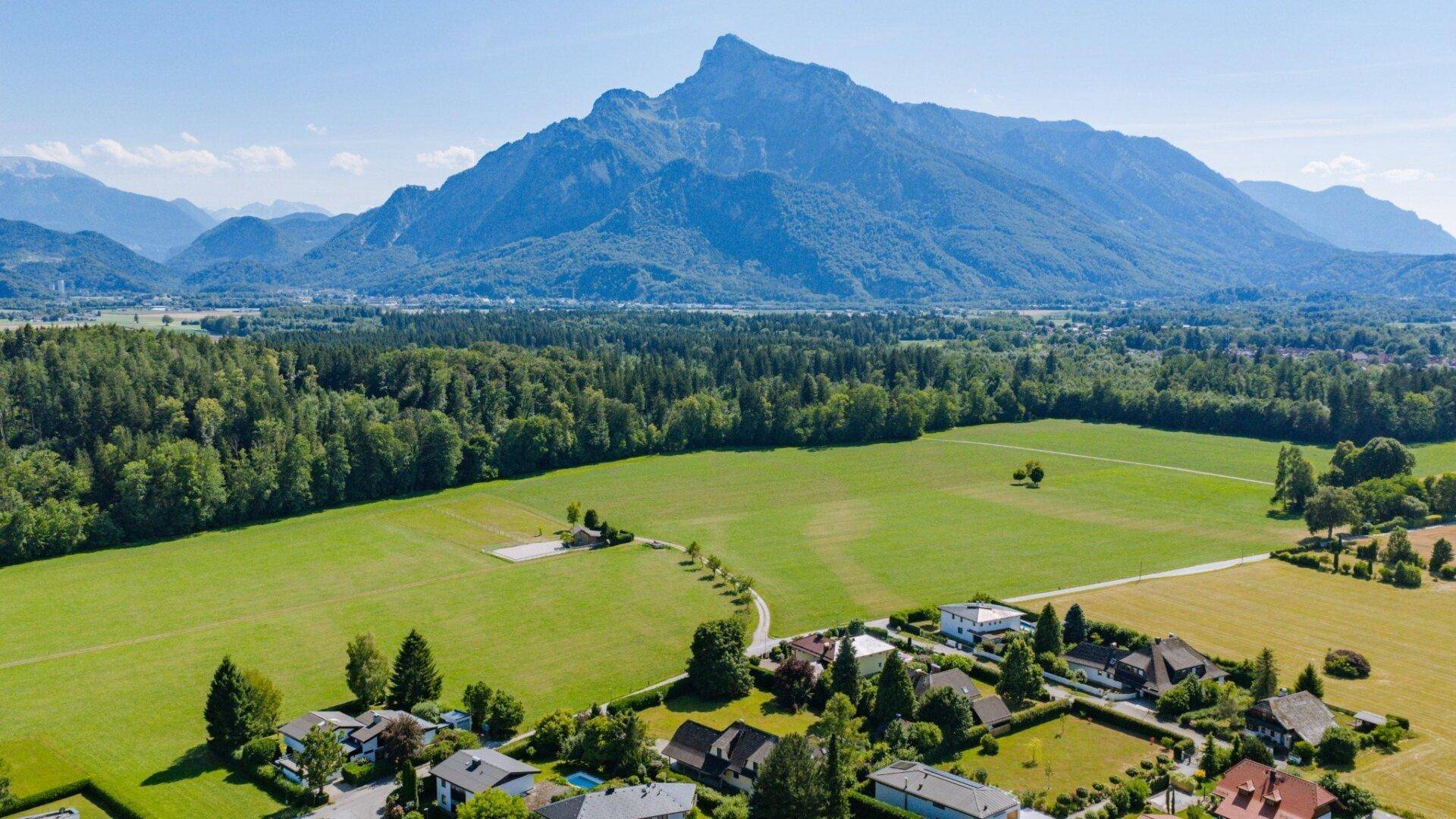 Weitläufige Luftaufnahme einer Wohnsiedlung mit grünen Feldern und imposanten Bergen im Hintergrund unter blauem Himmel.
