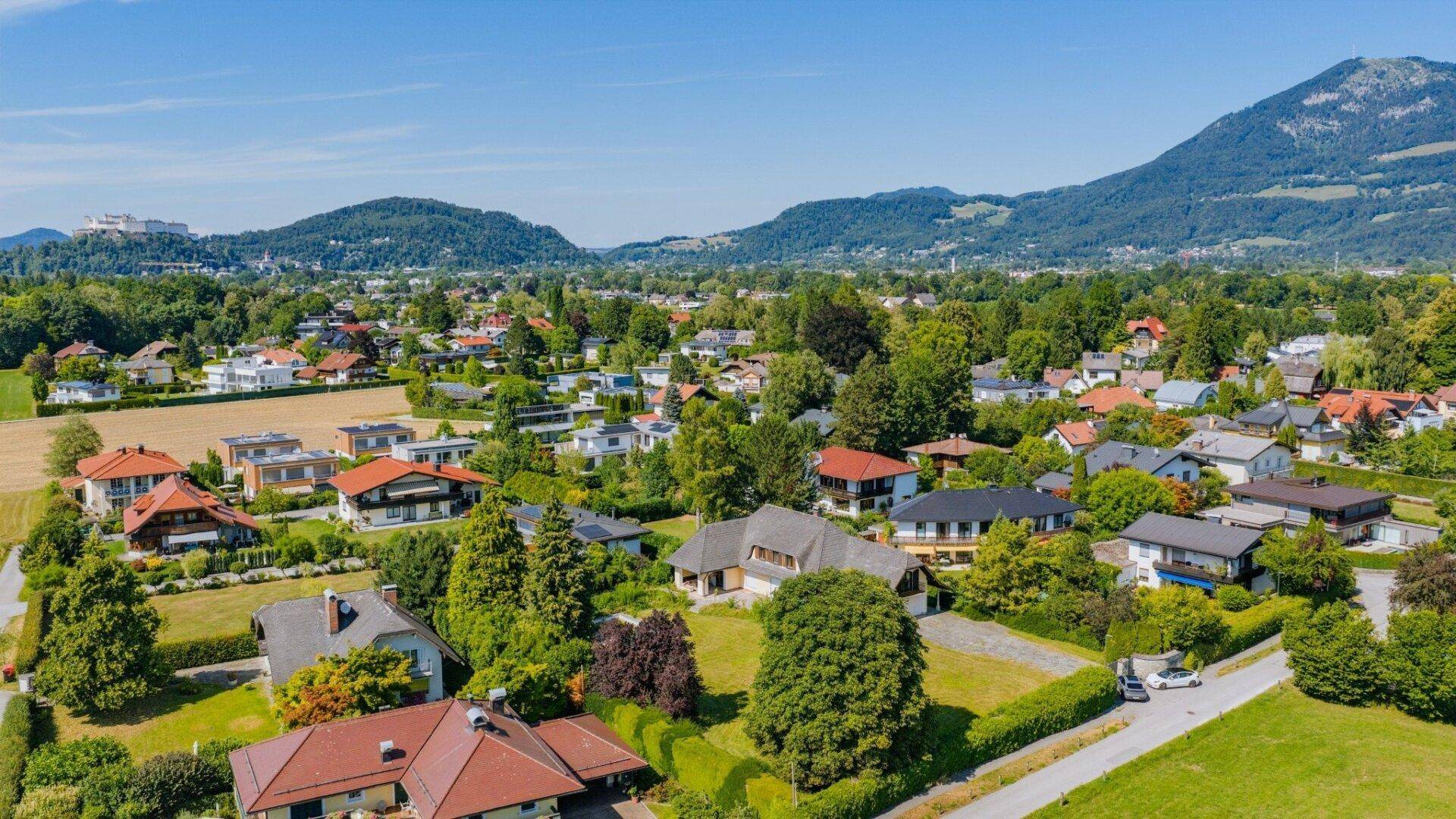 Luftaufnahme einer Wohngegend mit Einfamilienhäusern, umgeben von viel Grün und einem historischen Bauwerk in der Ferne.