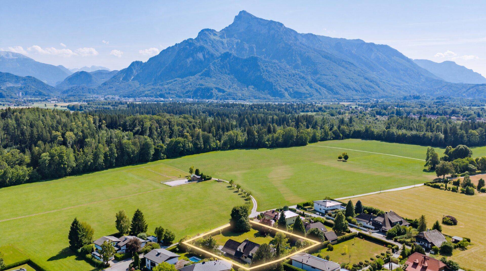 Luftaufnahme eines Grundstücks mit hervorgehobener Grenze, umgeben von Häusern und grüner Landschaft mit Bergen.