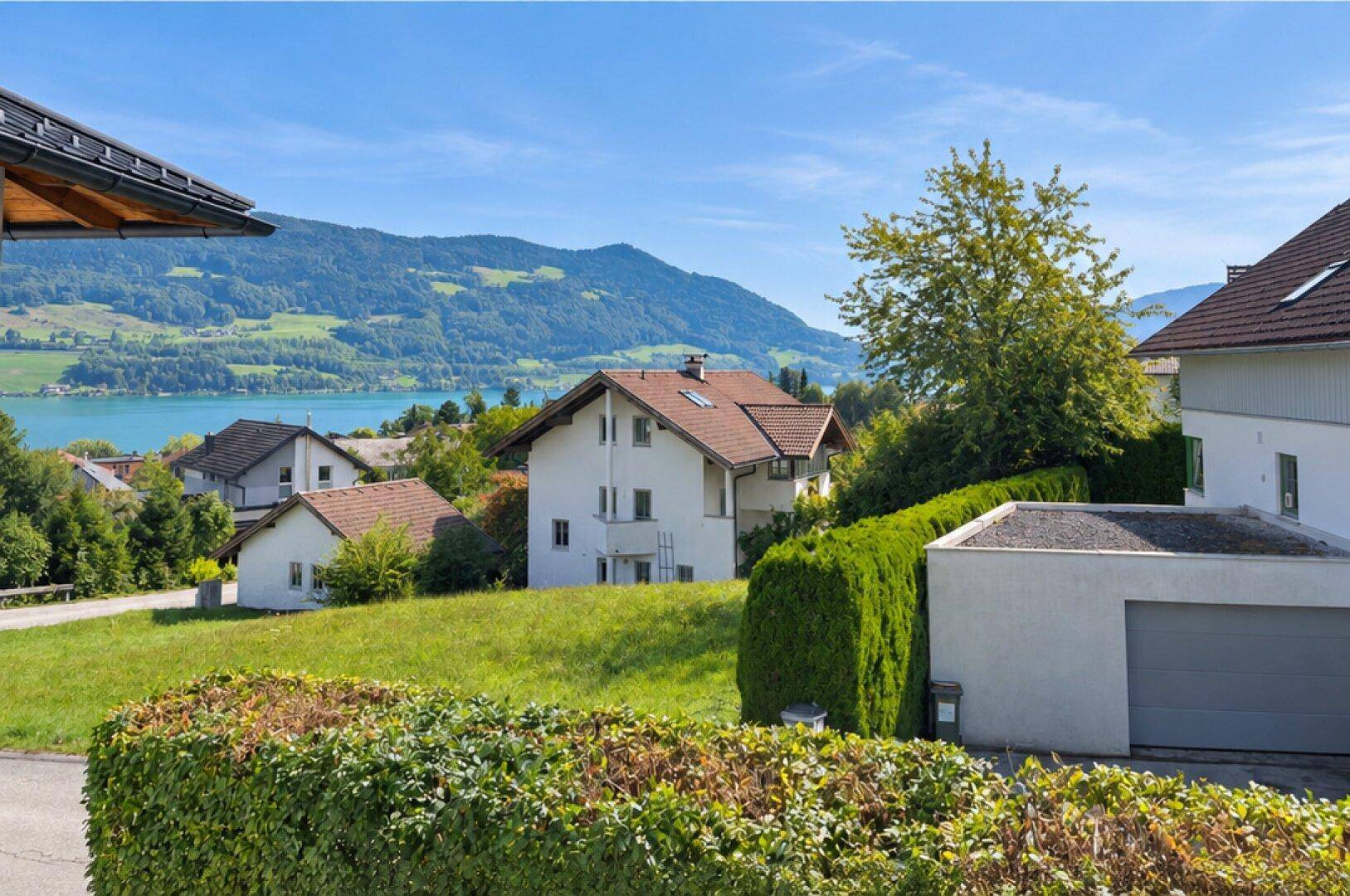 Malerische Aussicht auf Wohnhäuser, den See und die umliegende Berglandschaft unter blauem Himmel.