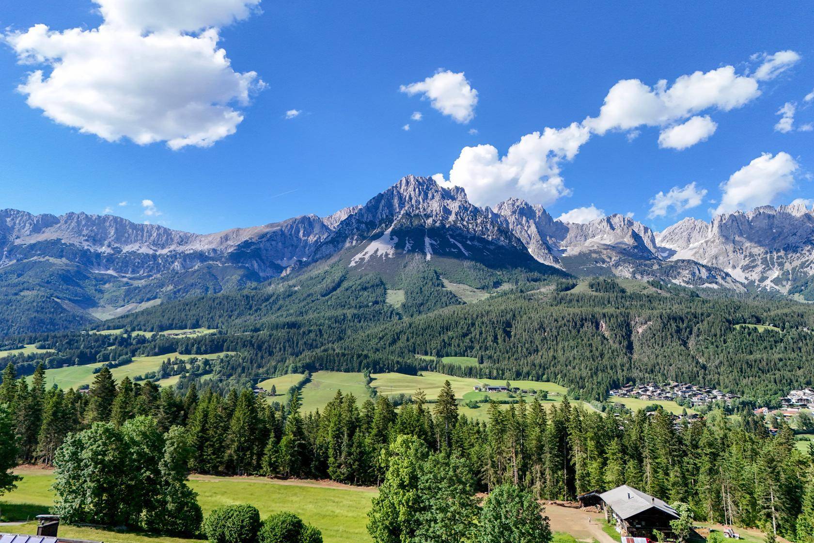 Weitläufige Landschaftsaufnahme mit imposanten Bergen, dichten Wäldern und grünen Tälern unter blauem Himmel.