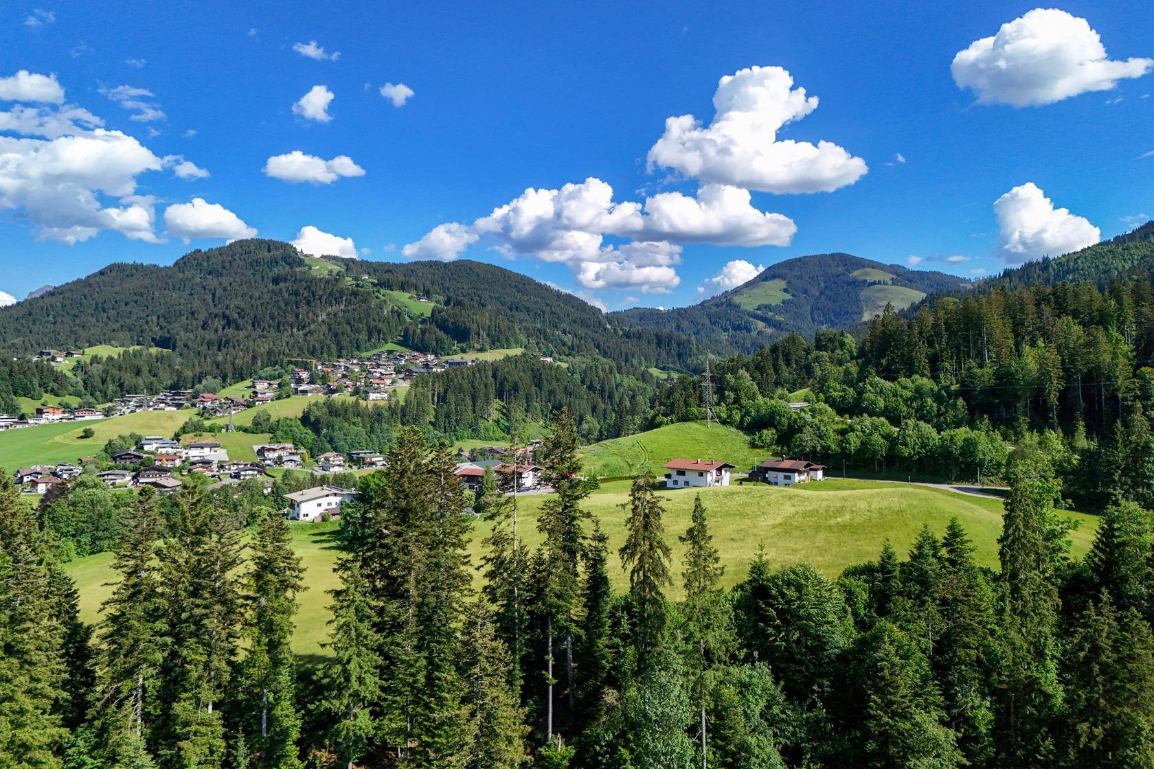 Blick auf ein grünes Tal mit verstreuten Häusern, bewaldeten Hügeln und einem strahlend blauen Himmel.