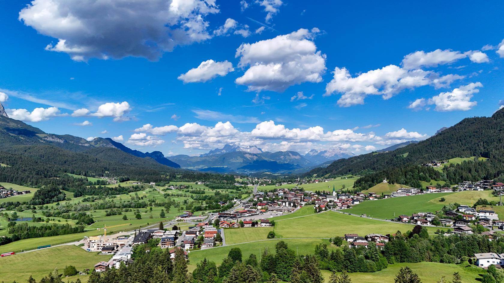 Panoramablick über ein malerisches Tal mit einem Dorf, umgeben von grünen Hügeln und fernen Bergen.