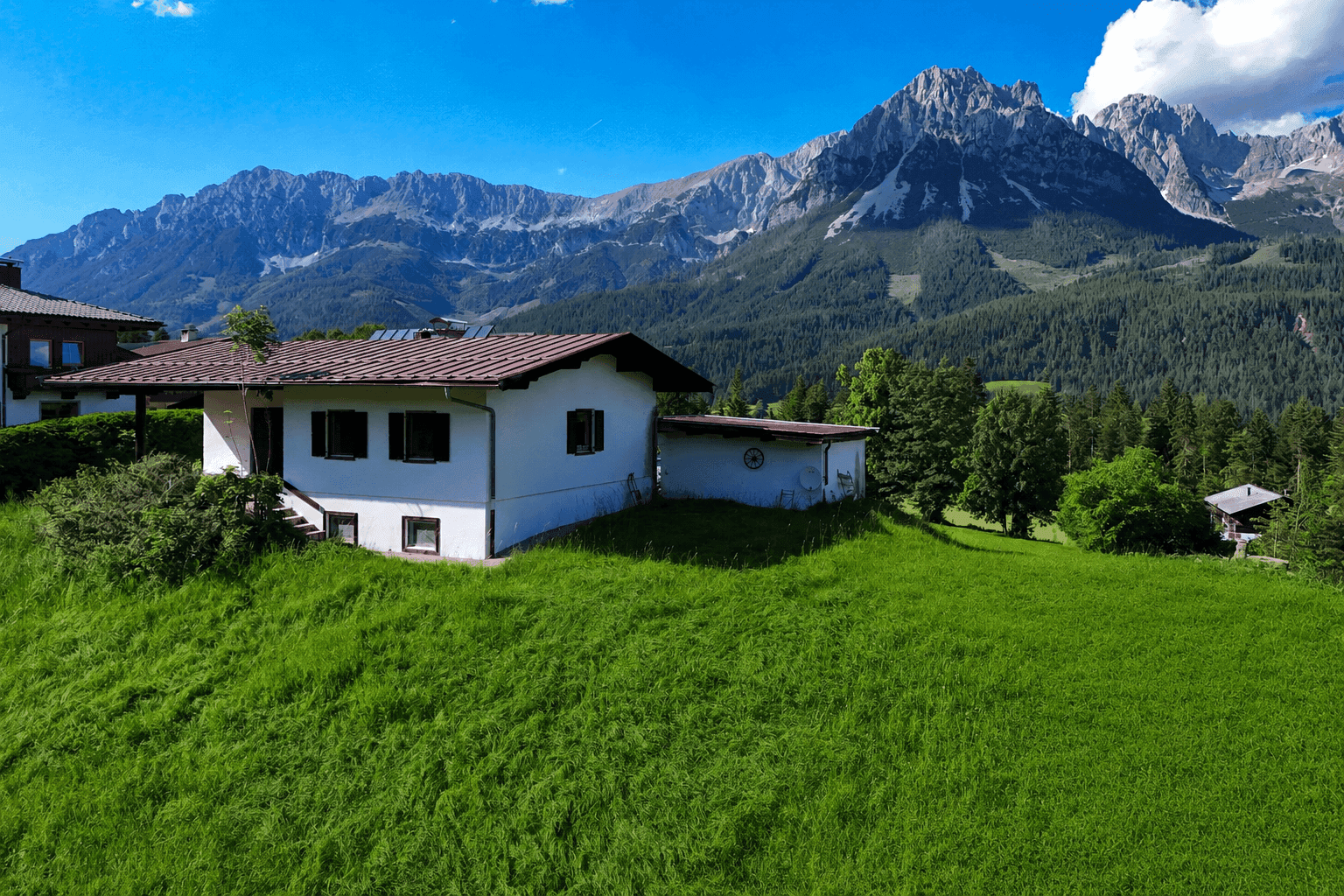Ein weißes Haus mit braunem Dach auf einem grünen Hügel vor einer beeindruckenden Bergkette.