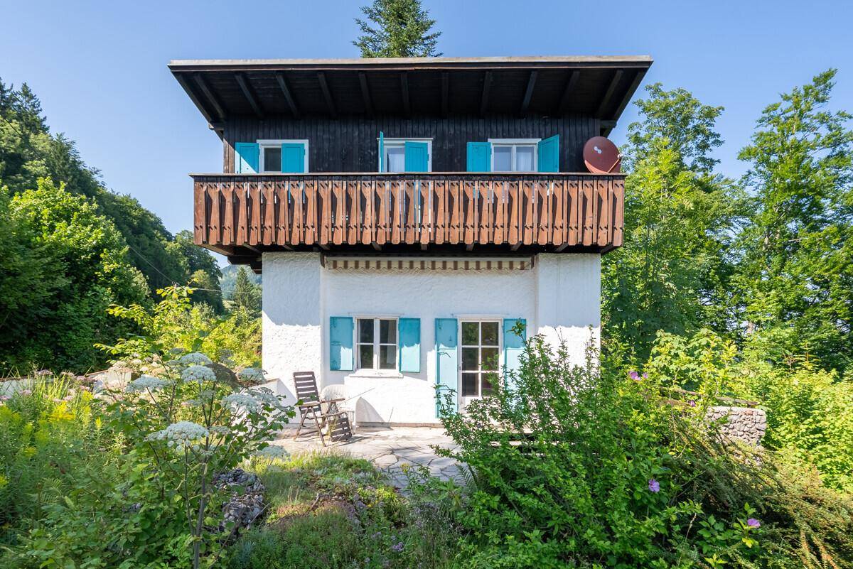 Frontansicht des Hauses mit blauen Fensterläden, Holzbalkon und einer kleinen Terrasse mit Sitzgelegenheiten.