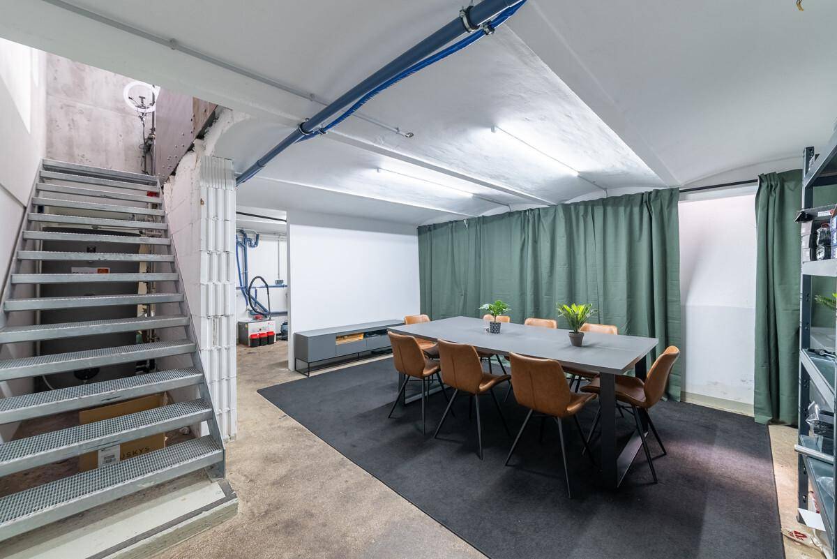 Kellerraum mit Metalltreppe, langem Tisch und Stühlen, ideal für Besprechungen oder als Aufenthaltsraum.