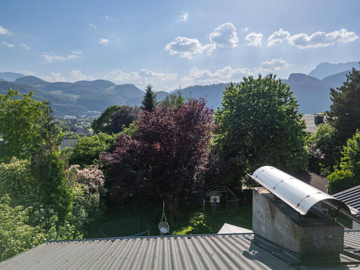 Weitblick über grüne Baumwipfel und Dächer auf die beeindruckende Berglandschaft bei Sonnenschein.