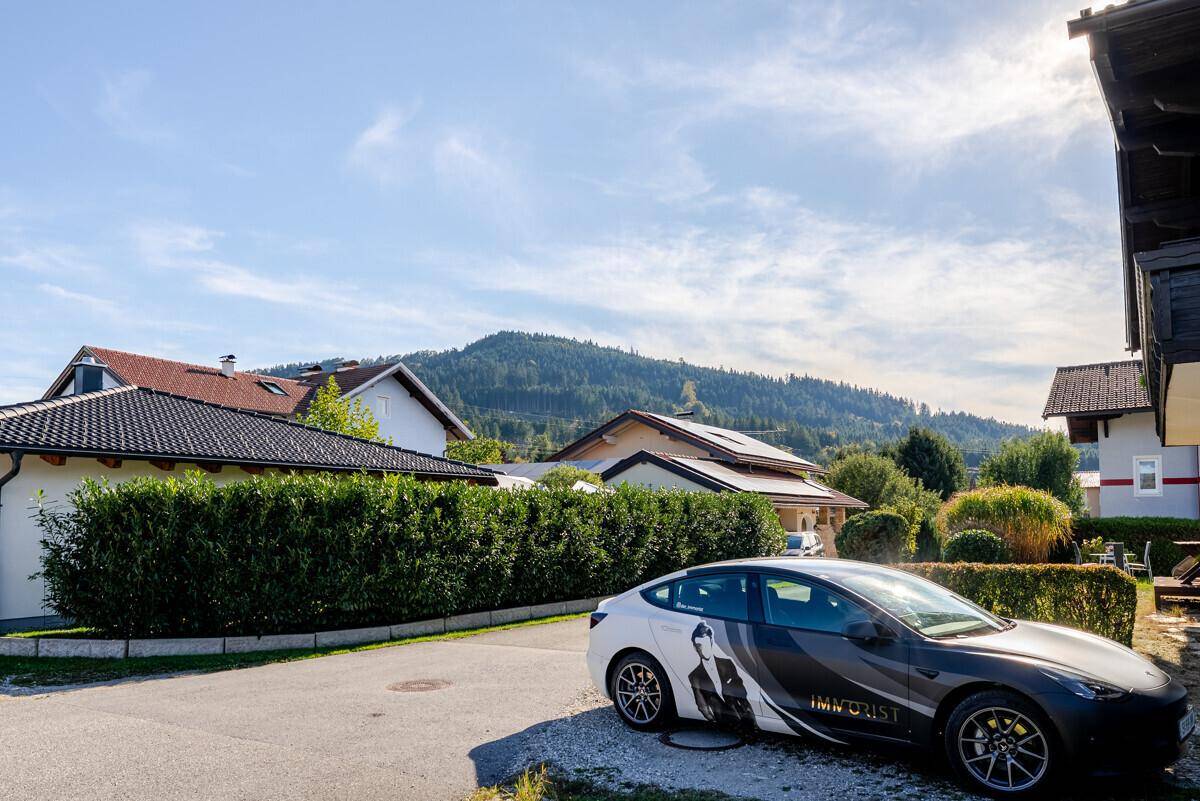 Straßenansicht mit geparktem Auto, Häusern, Hecke und einem bewaldeten Berg im Hintergrund.