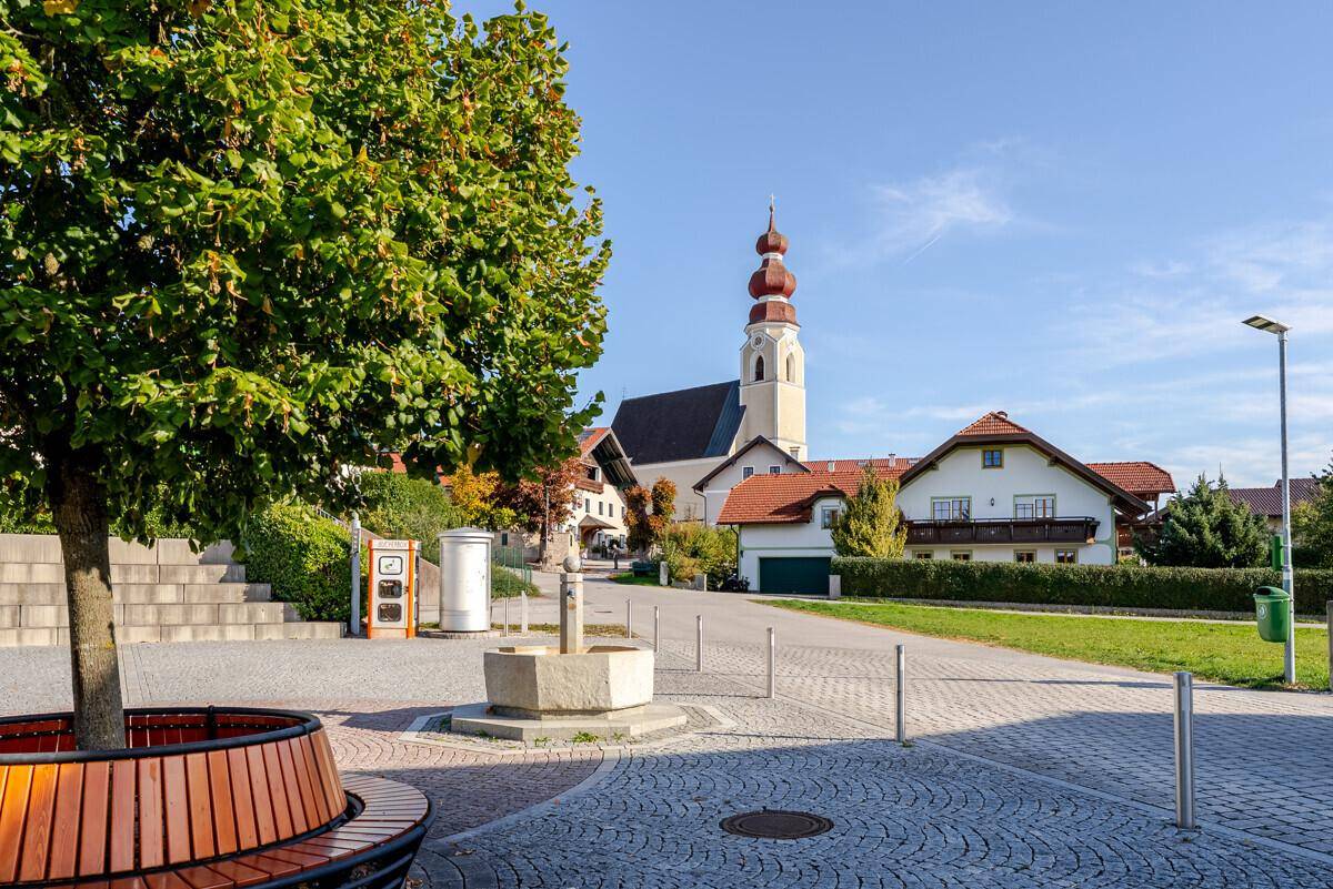 Dorfplatz mit Baum, Sitzbank, Brunnen und einer Kirche mit markantem Kirchturm im Hintergrund.