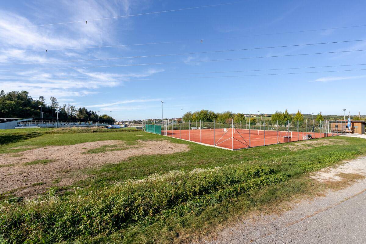 Roter Tennisplatz im Freien, umgeben von Grasflächen und einigen entfernten Gebäuden.