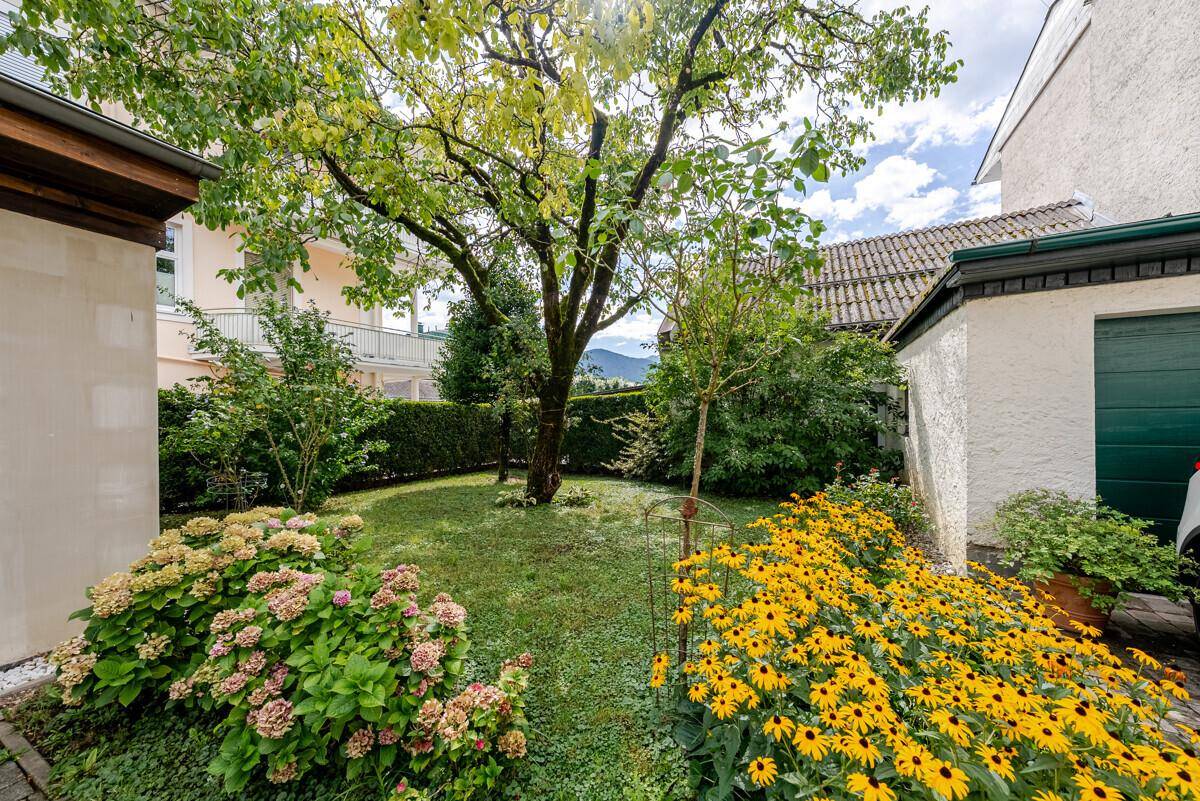 Gepflegter Gartenbereich mit Rasenfläche, blühenden Blumenbeeten und einem großen Baum.