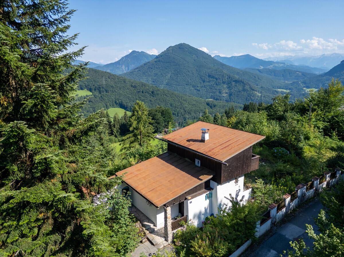 Luftaufnahme des Hauses mit braunem Dach, umgeben von dichten Wäldern und einer beeindruckenden Bergkulisse.