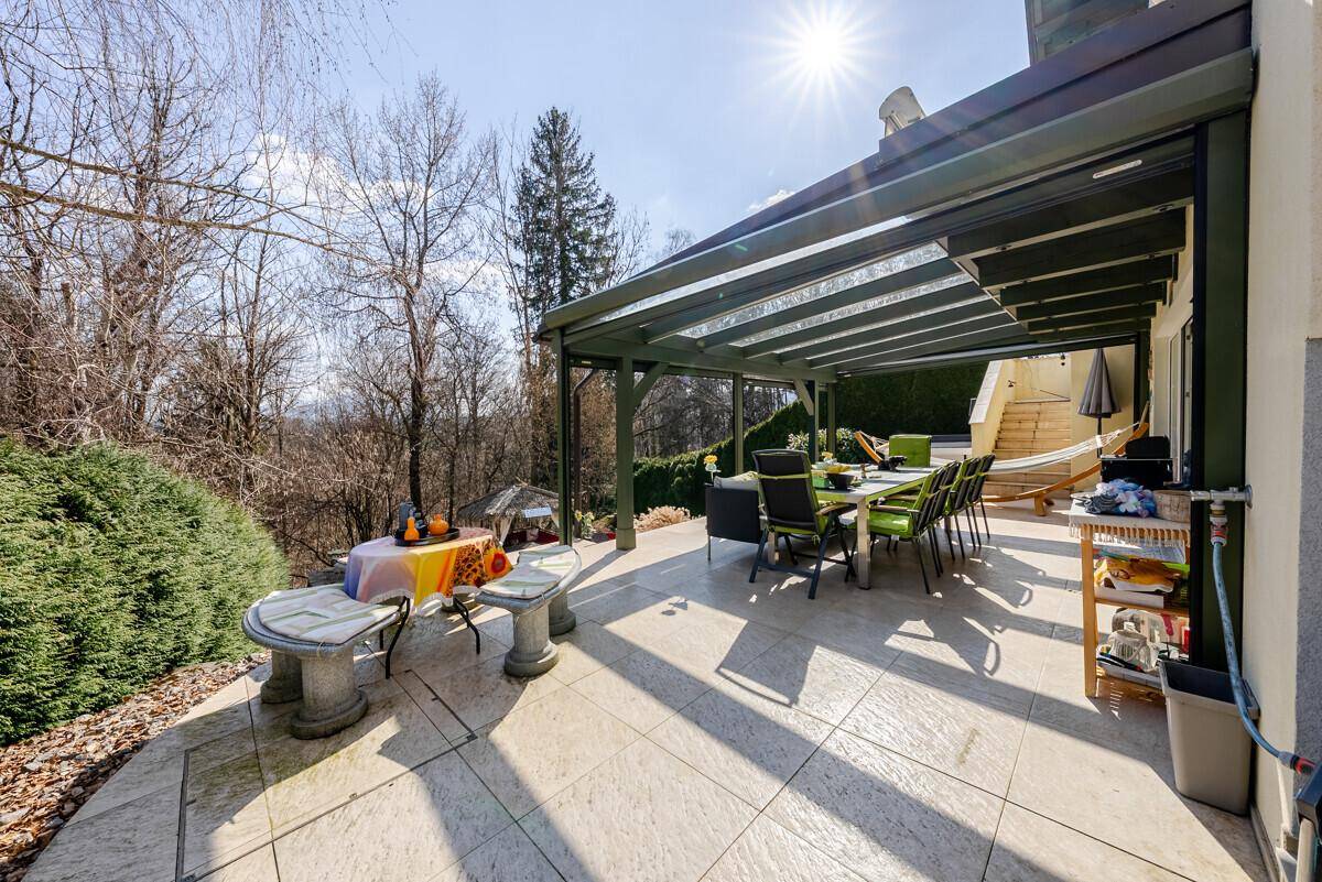 Sonnige Terrasse mit Essbereich und Steintischen, umgeben von Bäumen und Sträuchern.