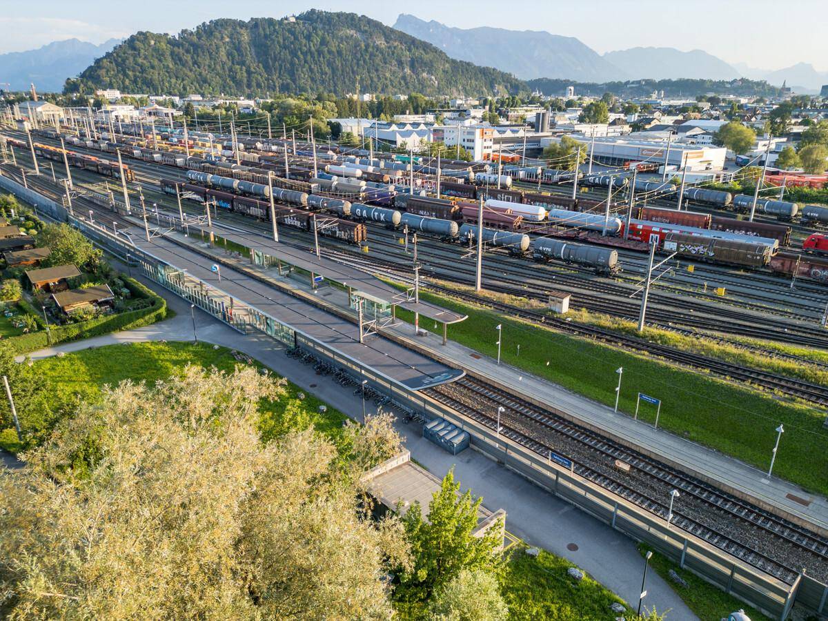 Luftaufnahme eines Bahnhofs mit zahlreichen Gleisen und Zügen vor einer Bergkulisse.
