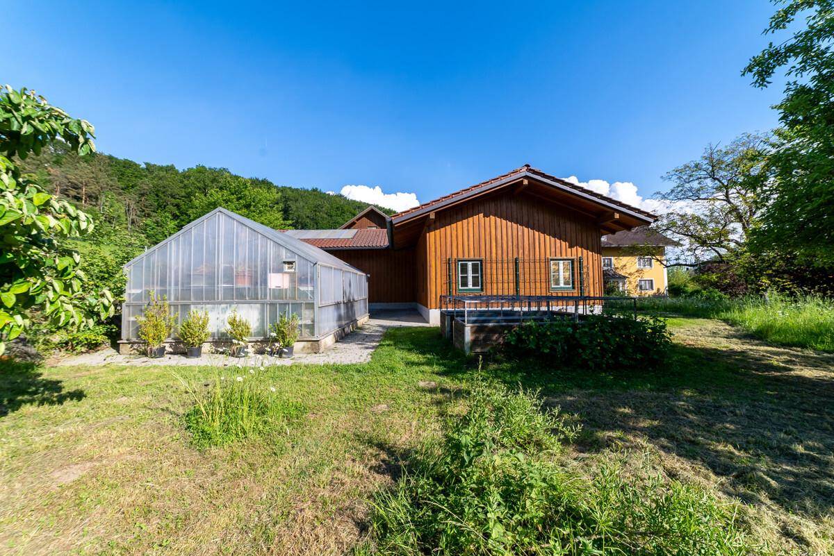 Holzverkleidetes Gebäude neben einem großen Gewächshaus, umgeben von Rasen und Bäumen.