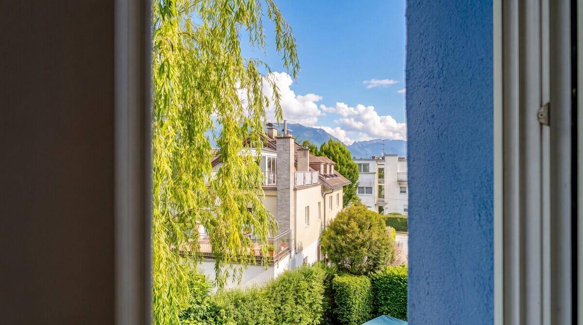 Blick aus dem Fenster auf grüne Bäume, umliegende Gebäude und ferne Berge unter blauem Himmel.