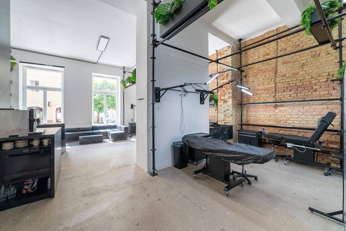 Behandlungsbereich in einem modernen Studio mit Liege, industriellen Regalen und Blick auf den Wartebereich.