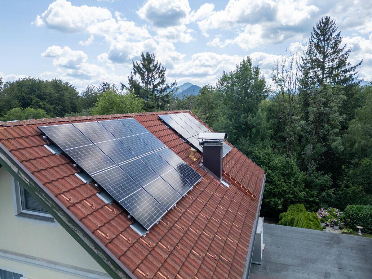 Nahaufnahme eines roten Ziegeldaches mit installierten Photovoltaikmodulen und bewaldeter Umgebung.