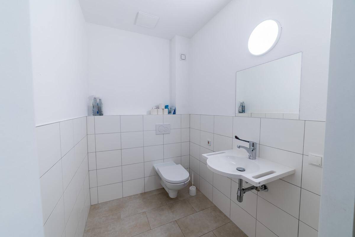 Sauberes WC mit wandhängender Toilette, Waschbecken, Spiegel und weißen Fliesen an Wänden und Boden.