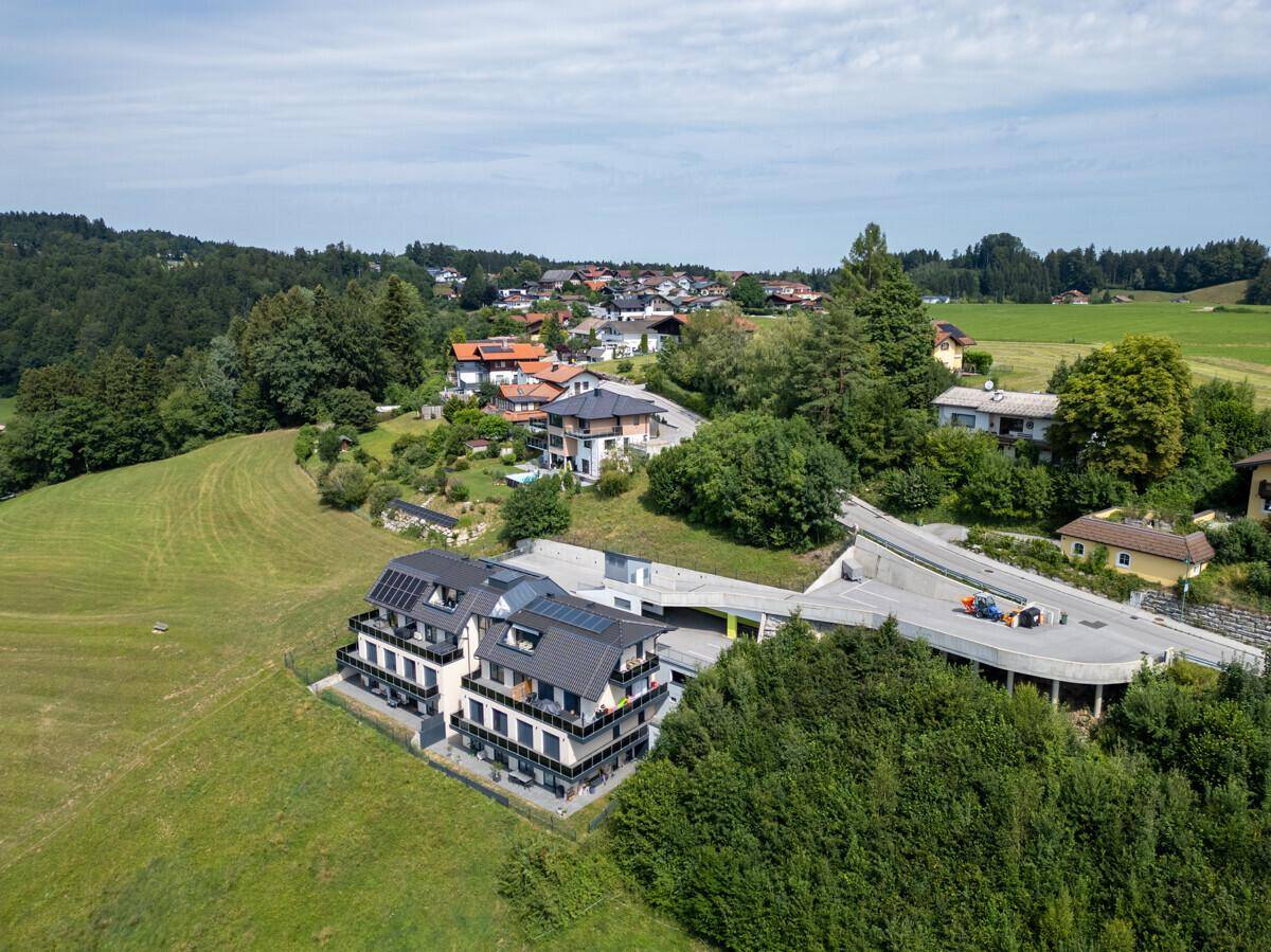 Luftaufnahme eines modernen Neubaus mit Solaranlagen, eingebettet in grüne Hügel und umgeben von weiteren Häusern.
