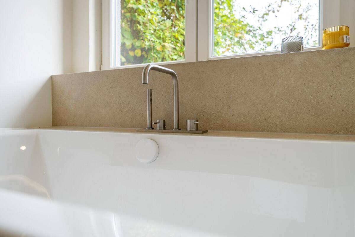Nahaufnahme einer modernen Badewanne mit Armaturen unter einem Fenster, das viel Licht hereinlässt.