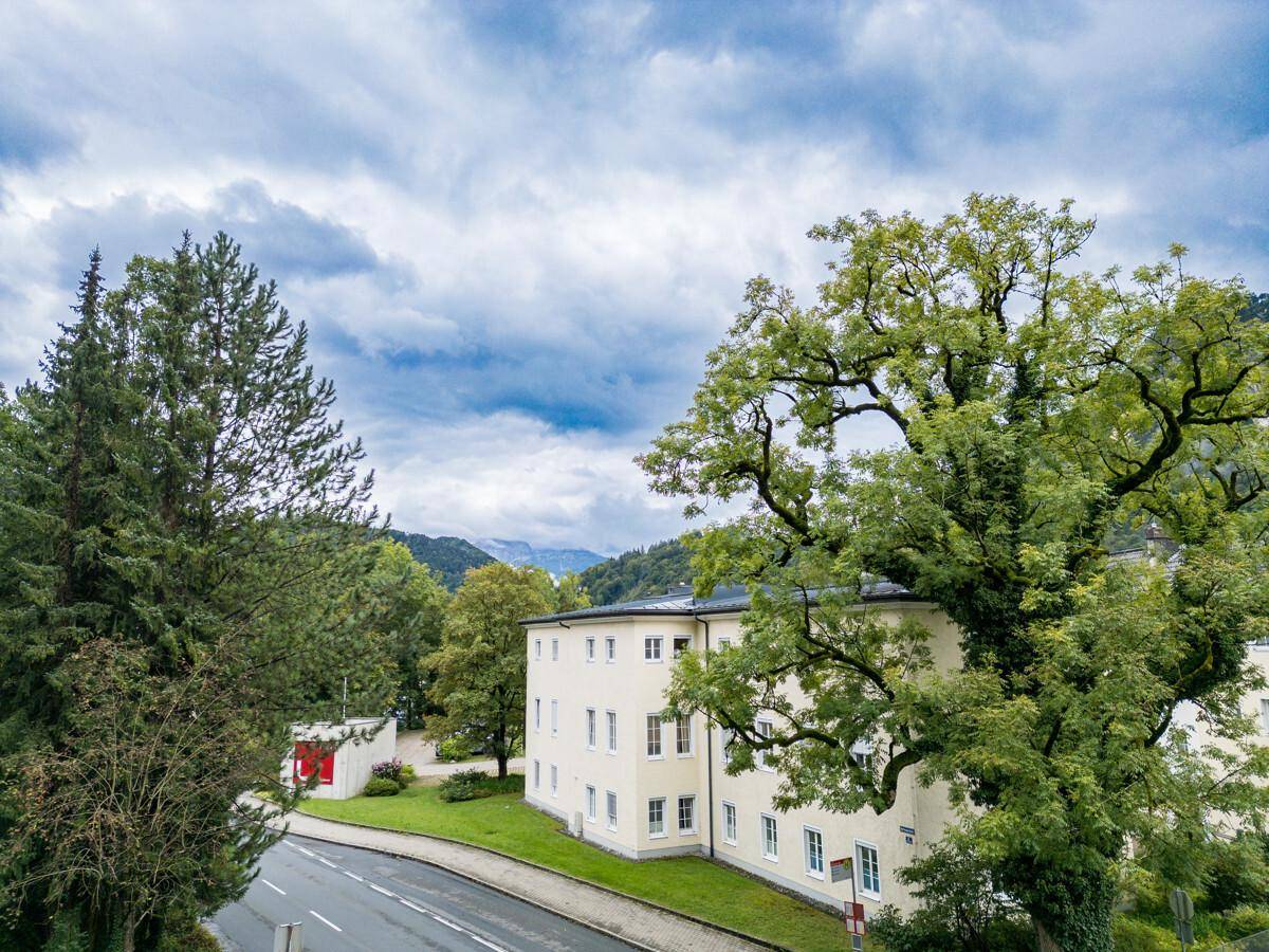 Historisches Gebäude umgeben von Bäumen und einer Straße mit Berglandschaft im Hintergrund.