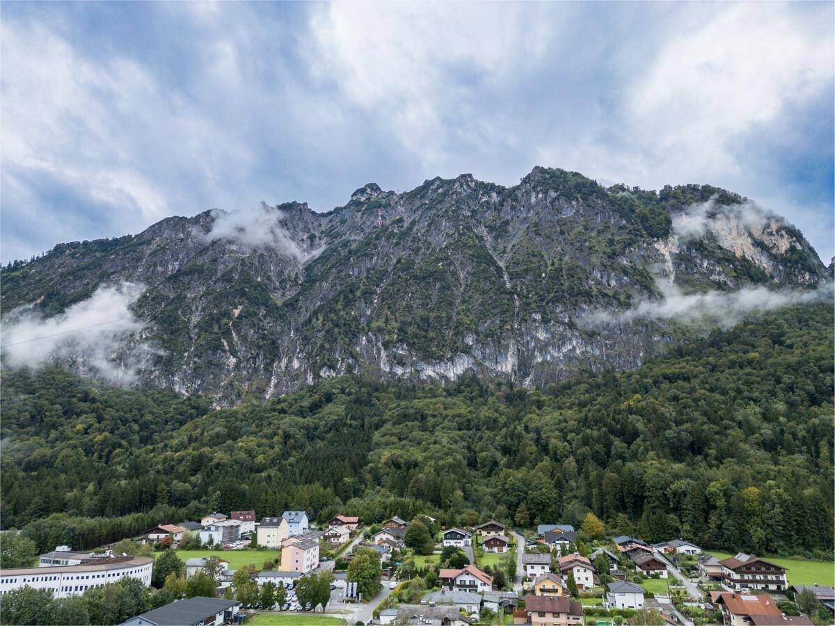 Weitläufige Luftaufnahme eines Dorfes, eingebettet in eine grüne Landschaft vor einer imposanten Bergkulisse.