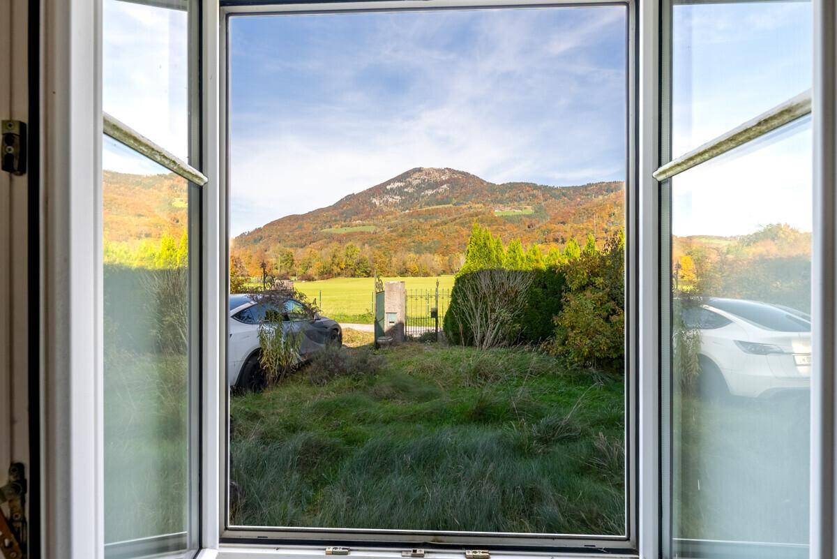 Offenes Fenster bietet einen weiten Blick auf eine grüne Wiese, Berge und herbstliche Bäume.
