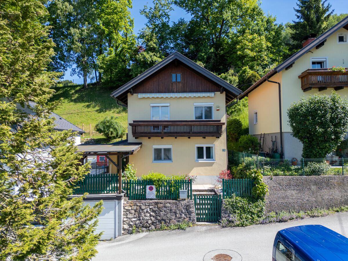 Charmantes Einfamilienhaus mit Balkon und Carport, umgeben von viel Grün und Bäumen.