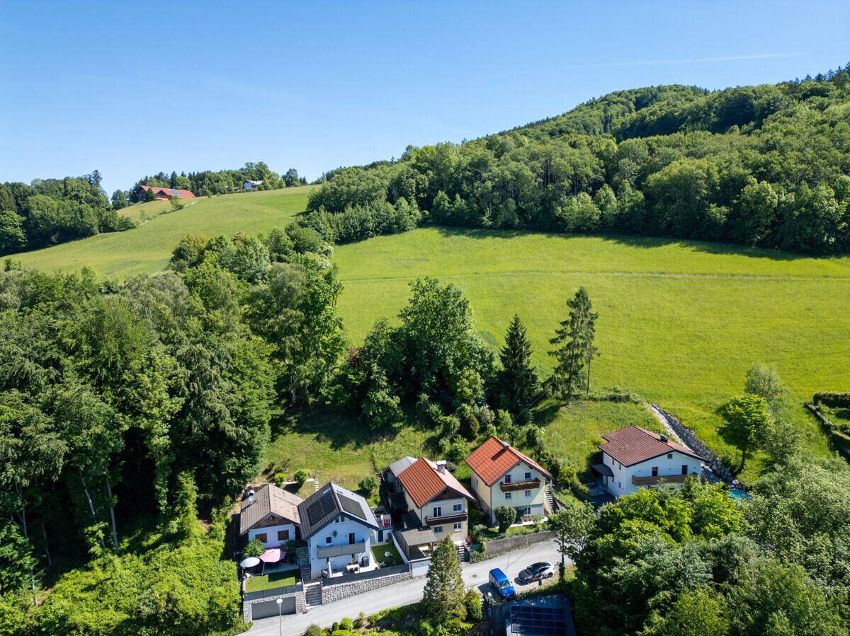 Luftaufnahme einer Wohnsiedlung am Hang, umgeben von Wäldern und grünen Wiesen mit Bergen im Hintergrund.