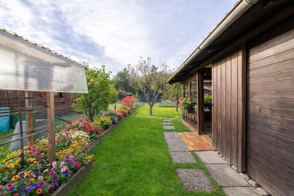 Seitenansicht eines Holzhauses mit gepflegtem Gartenweg, Blumenbeet und Gewächshaus im Hintergrund.