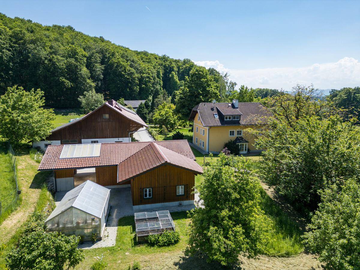 Luftaufnahme des Anwesens mit mehreren Gebäuden, Gewächshaus und umgebender grüner Landschaft.