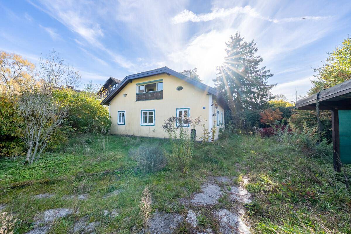 Gelbes Haus mit dunklem Dach und einem verwilderten Garten unter strahlendem Sonnenschein.