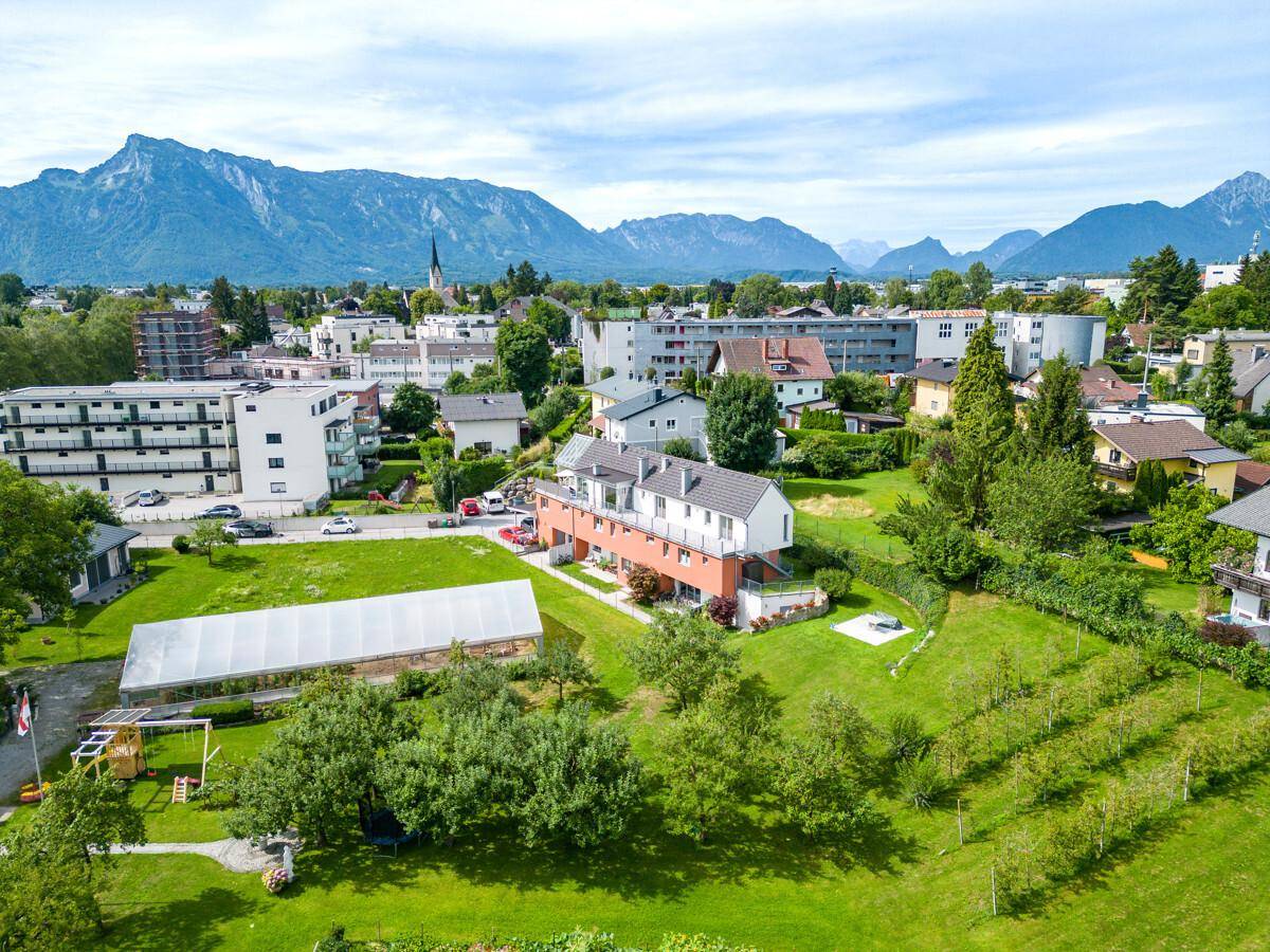 Luftaufnahme einer Wohnsiedlung mit viel Grün, Gewächshaus und majestätischem Bergpanorama.