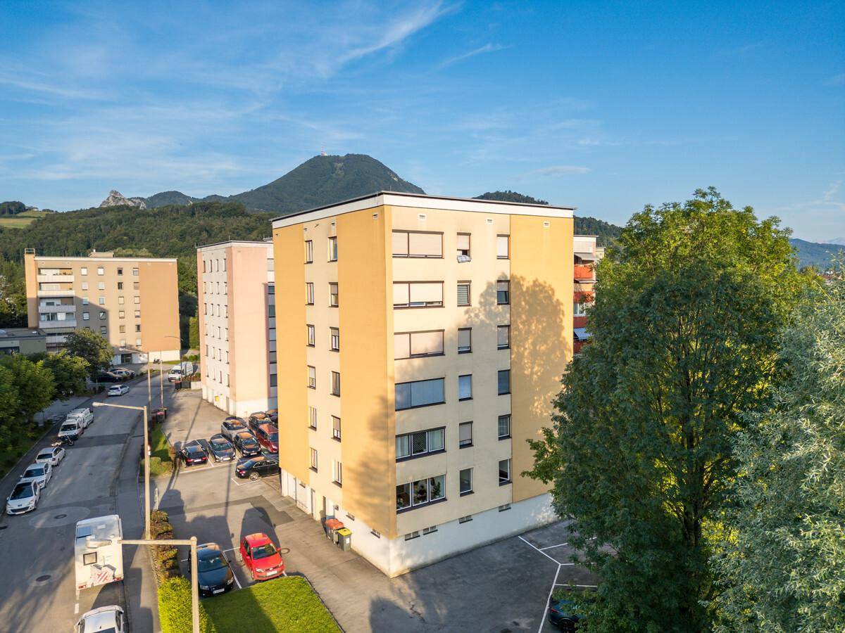 Mehrfamilienhaus in einer Wohnsiedlung mit Parkplätzen und Bergblick im Hintergrund.