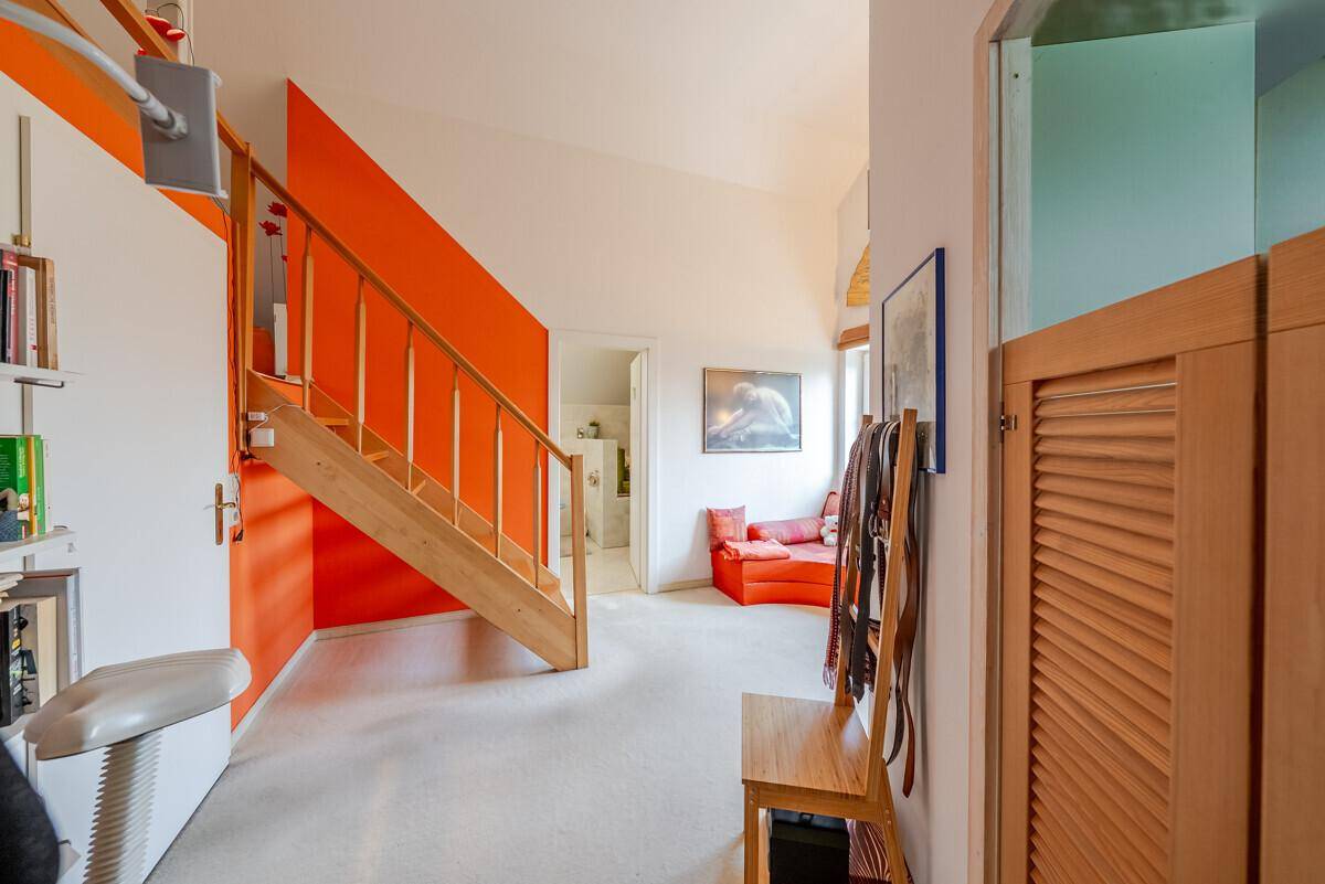 Heller Flur mit Holztreppe, orangefarbener Akzentwand und Zugang zu weiteren Räumen.