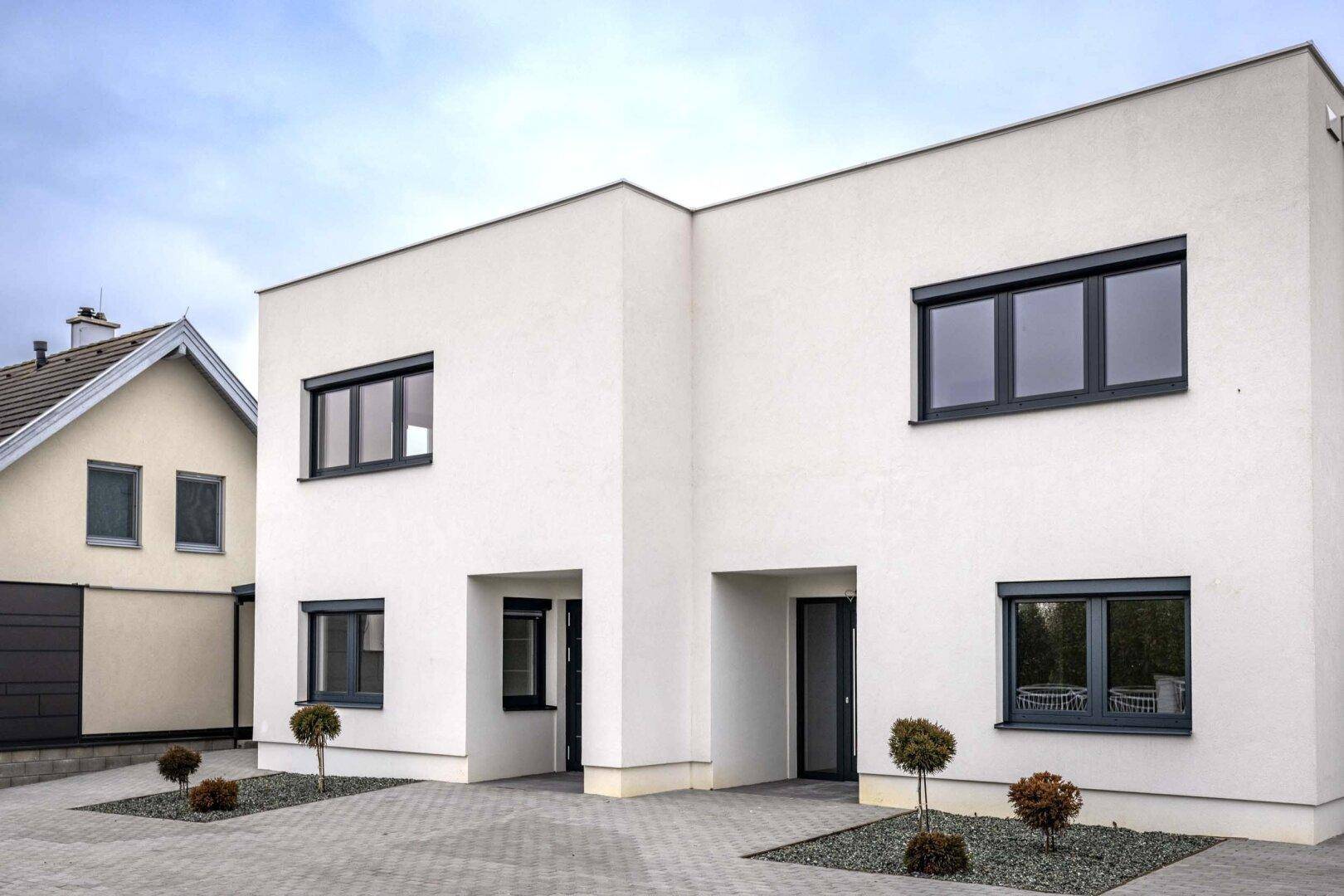 Moderne Doppelhaushälfte mit weißer Fassade, dunklen Fenstern und gepflasterter Einfahrt.