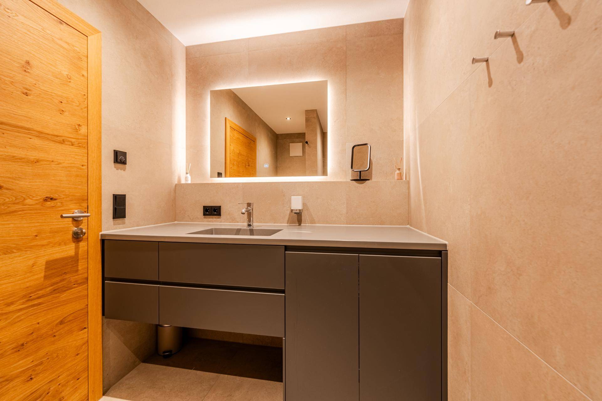 Modernes Badezimmer mit Einzelwaschbecken, beleuchtetem Spiegel und großformatigen Fliesen.
